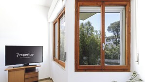 Ein Zimmer mit Fernseher auf einem Holztisch, ein großes Fenster mit Holzrahmen und Blick auf Grünflächen und Gebäude.