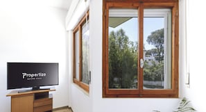 Ein Zimmer mit Fernseher auf einem Holztisch, ein großes Fenster mit Holzrahmen und Blick auf Grünflächen und Gebäude.
