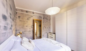 A bedroom with a bed, a nightstand, a wardrobe, a hanging light fixture, and a door leading to another room. - 3BR Family Apartment by Propertize (Cagliari)