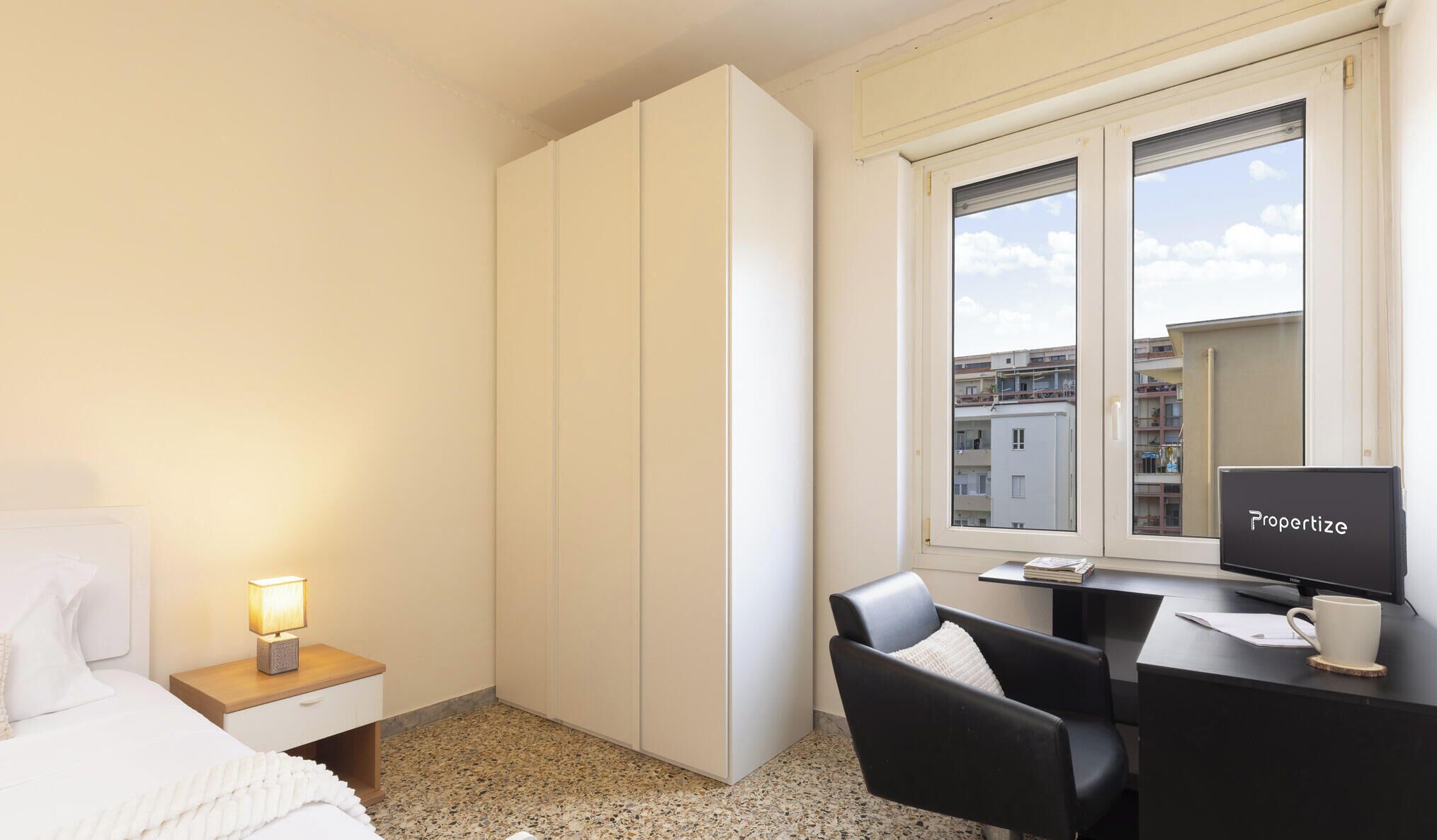 A hotel room with a bed, bedside table with a lamp, a desk with a computer, a chair, and a window with a view of buildings.