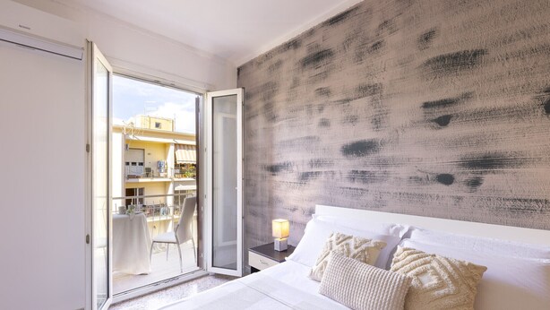 Un dormitorio con cama, mesita de noche, lámpara y vistas a un balcón con mobiliario exterior.