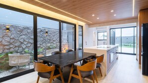 A modern kitchen with a large island, a dining area with a black table and wooden chairs, and large windows offering a view of a stone wall and greenery. - Modern House: 3 Suites | Jacuzzi | 450m from Beach (Florianópolis)
