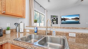 Una cocina con encimeras de granito, lavamanos de acero inoxidable, armarios de madera, un ventanal con cortinas, un televisor de pantalla plana montado y un enchufe.