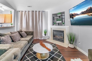 A living room with a sofa, a round coffee table, a fireplace, a flat-screen TV, and a view of mountains.