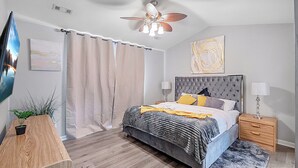Un dormitorio con una cama, un ventilador de techo, una mesita de noche, una lámpara, una planta en maceta y un cuadro en la pared.