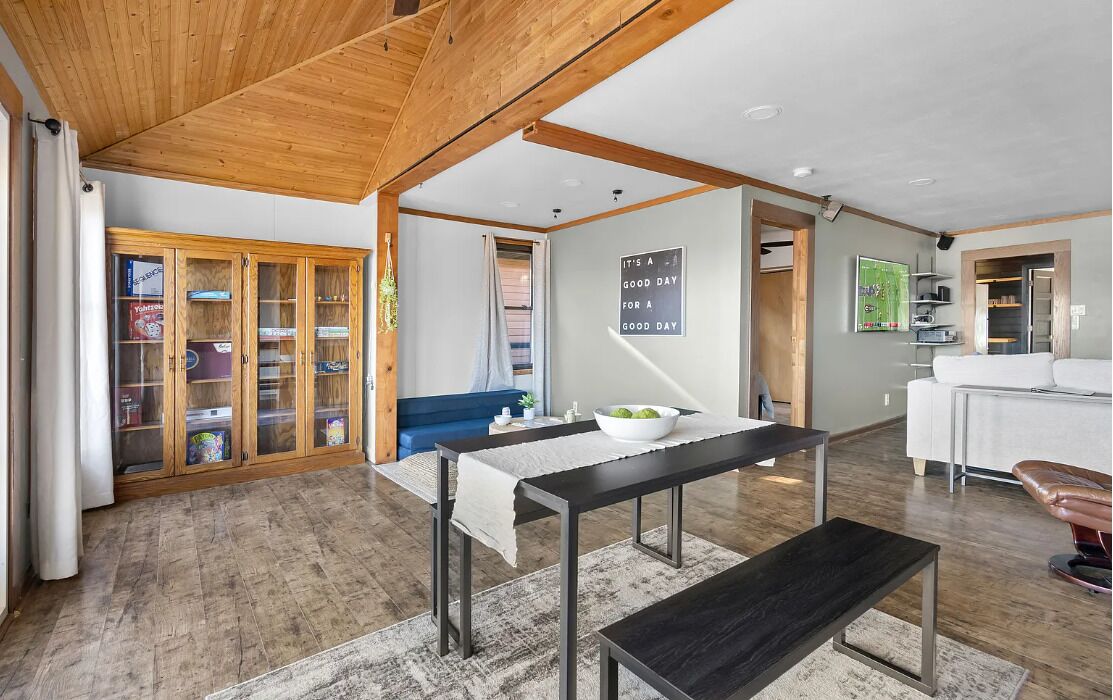 Un vaste espace de vie doté d’un plafond en bois, d’une table à manger avec des bancs, d’un canapé bleu et d’un coin cuisine visible à travers une porte ouverte.