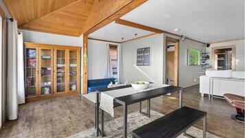 Un vaste espace de vie doté d’un plafond en bois, d’une table à manger avec des bancs, d’un canapé bleu et d’un coin cuisine visible à travers une porte ouverte.