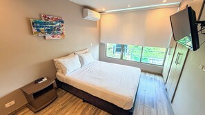 A modern bedroom with a large bed, a nightstand, a TV, and a window with a view of greenery. - Santorini Apartamentos amoblados (Pereira)