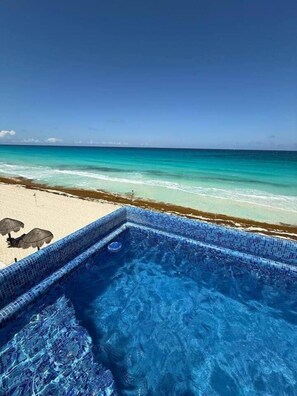 Visão nítida de uma piscina com borda azulejada, uma praia arenosa e o oceano ao fundo.