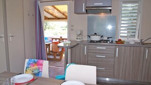 Une cuisine compacte équipée d’une cuisinière à gaz, d’une machine à café et d’un évier. On aperçoit un coin repas avec une table et des chaises à travers une porte ouverte.