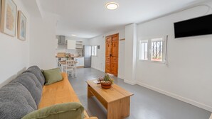 Una sala de estar con sofá, una mesa de centro de madera con una planta, un televisor montado en la pared y una cocina al fondo.