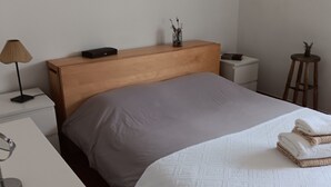 A neatly made bed with a grey blanket, a bedside lamp, a small bedside table with a book, a wooden nightstand, and a small stool with a plant. - Apartment 'Garro1' with Mountain Views (Donostia / San Sebastián)