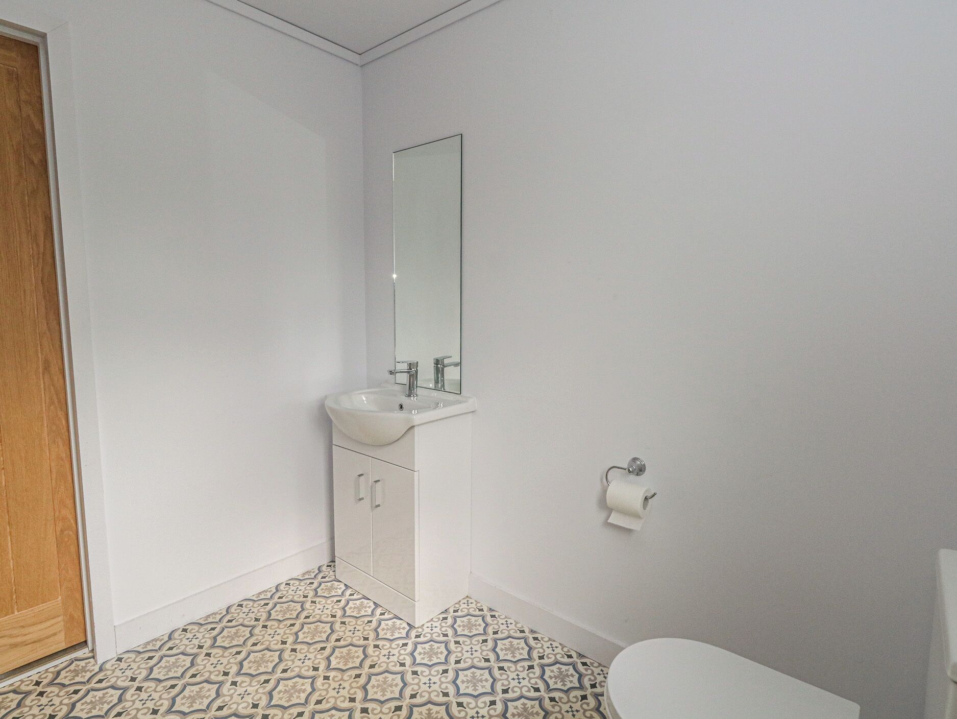 A bathroom with a white sink, mirror, toilet, patterned floor tiles, and a wooden door.