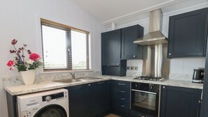 A compact kitchen with a built-in washer, oven, and stove, dark cabinetry, and a window with blinds. - Grass Roots (Templecombe)