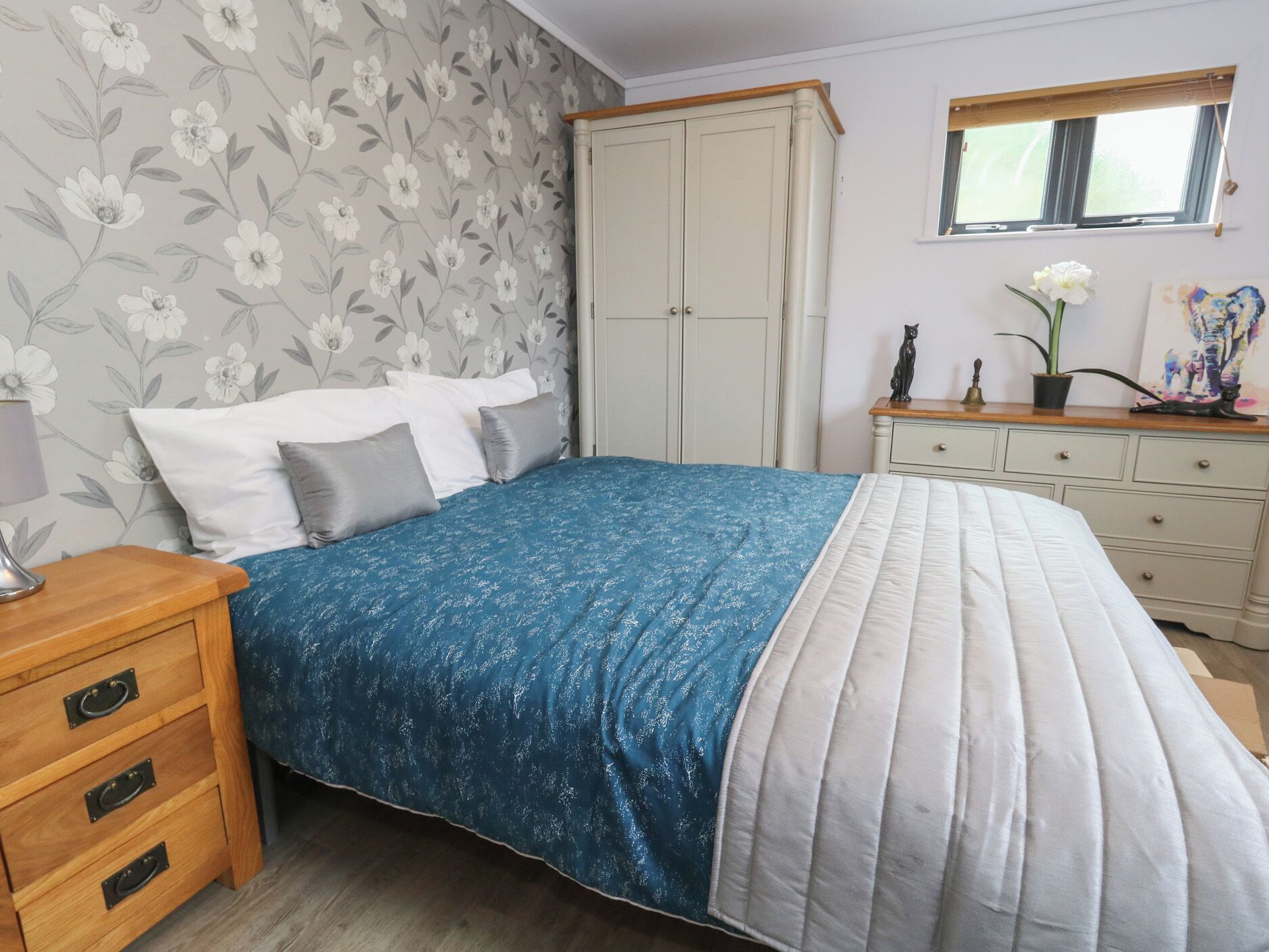 A bedroom with a bed, a nightstand, a dresser, a wardrobe, a window, and a floral wallpaper.