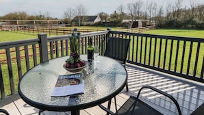 Ein runder Glastisch mit einer Zeitschrift, einer Schüssel mit Trauben und einer Flasche auf einer Terrasse mit Blick auf ein eingezäuntes Feld und Gebäude.