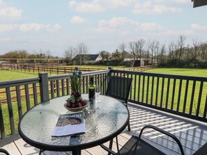 A round glass table with a magazine, a bowl of grapes, and a bottle on a deck with a view of a fenced field and buildings. - Grass Roots (Templecombe)