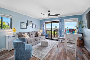 Ein Wohnzimmer mit grauem Sofa, blauem Sessel und einem Esstisch mit Stühlen. Es gibt einen Deckenventilator, ein Fenster mit Vorhängen und Blick ins Freie.