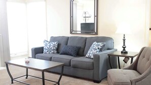 Ein Wohnzimmer mit grauem Sofa, gemusterten Kissen, einem Couchtisch, einem Beistelltisch mit Lampe und einem Sessel.
