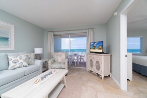 Ein Wohnzimmer mit einer Couch, einem Couchtisch mit Kerzen, einem Fernseher auf einer Kommode und Blick auf den Strand durch eine Glastür.