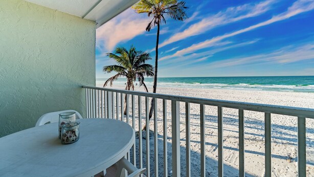 A balcony with a view of the beach, a table with chairs, and a palm tree. - Nautilus 209 by Nautilus Vacations - Beachfront Condo, Heated Pool, Racket Court (Holmes Beach)