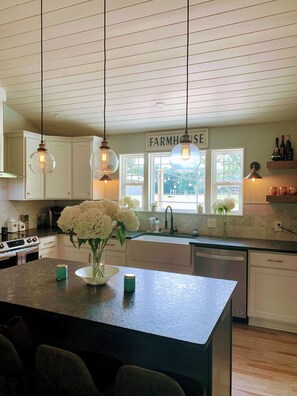Une cuisine avec des meubles blancs, un panneau décoratif de style rustique, des suspensions lumineuses et un vase rempli de fleurs posé sur l’îlot central.