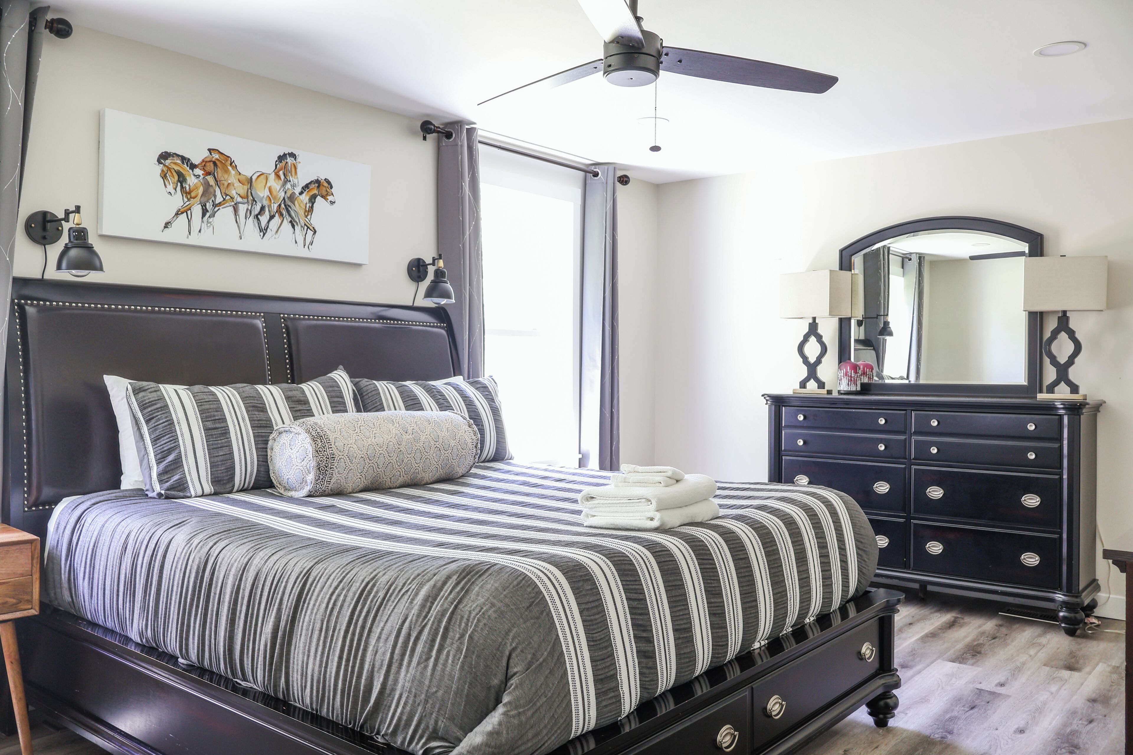 Un dormitorio con cama, ventilador de techo, cómoda, espejo y un cuadro en la pared.