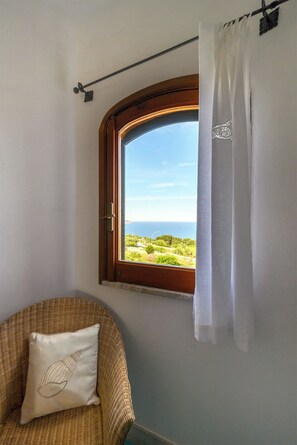 Ein Zimmer mit Fenster und Blick auf eine Landschaft, ein Korbstuhl mit Kissen sowie ein Vorhang.