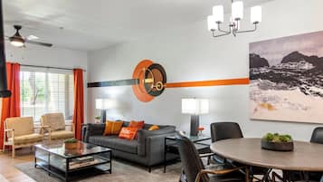 Un salon moderne avec un canapé, des fauteuils, une table basse et une table à manger. Il y a un tableau au mur et un ventilateur de plafond.