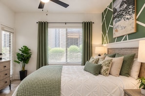 A bedroom with a bed, a nightstand, a lamp, a window with curtains, a ceiling fan, and a painting on the wall. - WestWorld + TPC | Heated Pool + Hot Tub (Scottsdale)