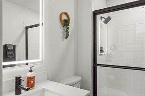A bathroom with a toilet, a sink with a soap dispenser, a mirror, and a shower area. - WestWorld + TPC | Heated Pool + Hot Tub (Scottsdale)