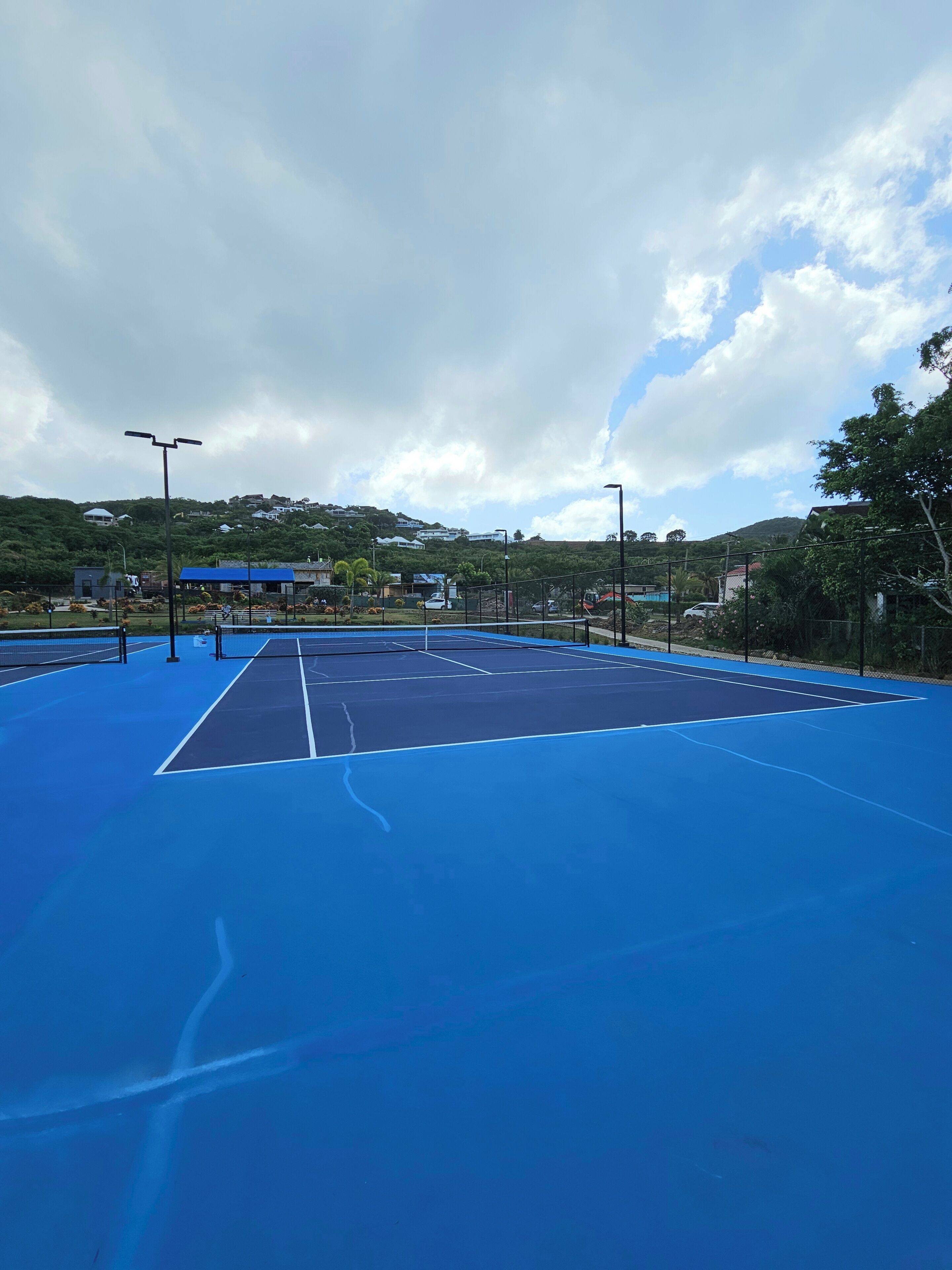 A tennis court with a blue surface, surrounded by a fence, and a cloudy sky above.
