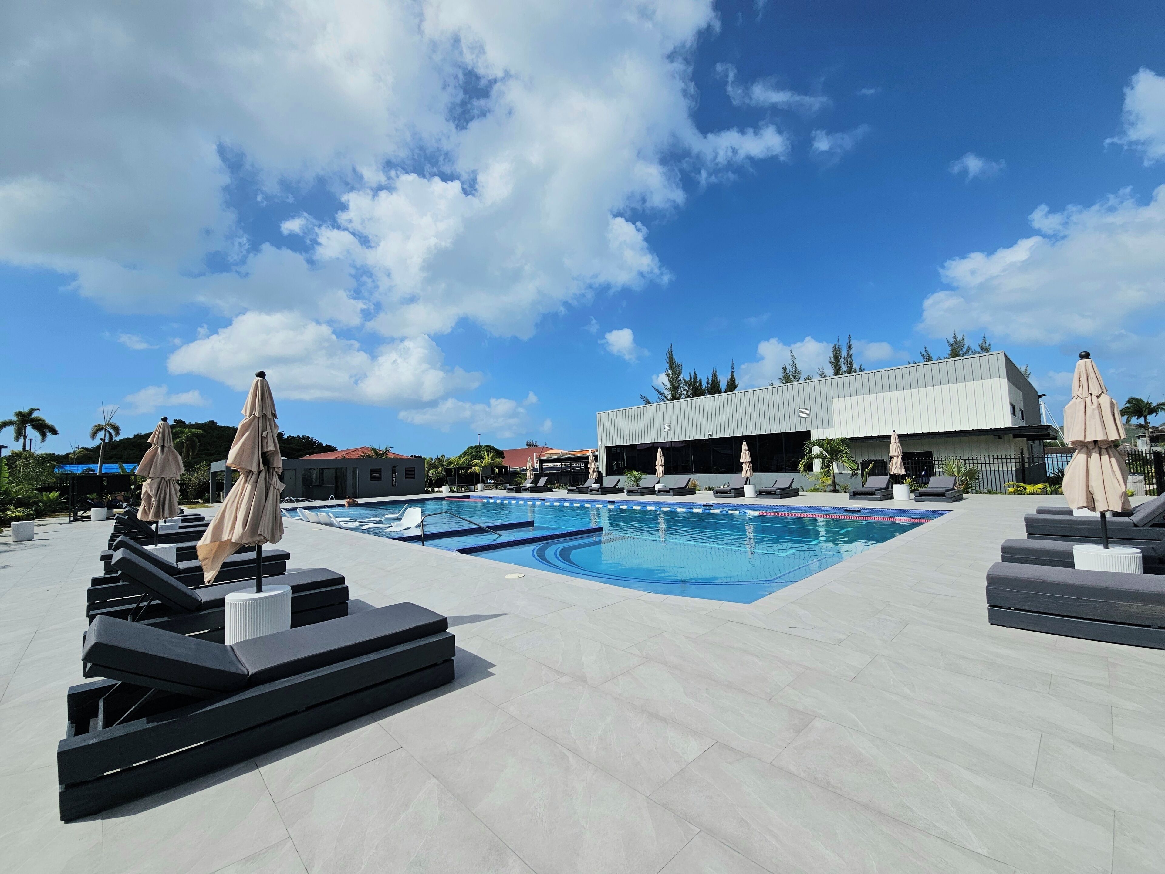 Zona de piscina con sillones de descanso y sombrillas, un edificio moderno y palmeras al fondo.