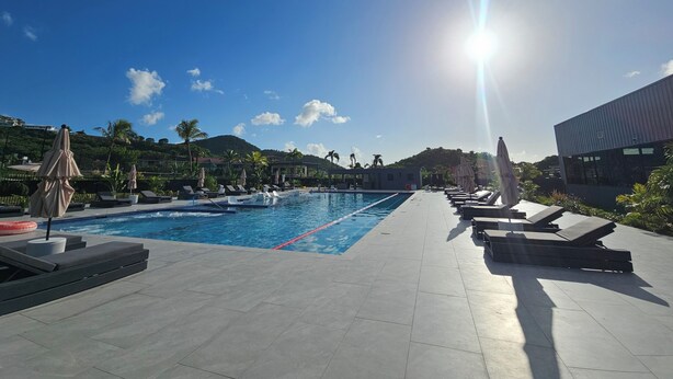 En swimmingpool med solstolar och parasoller, omgiven av ett vackert anlagt område med palmer och utsikt över kullar.