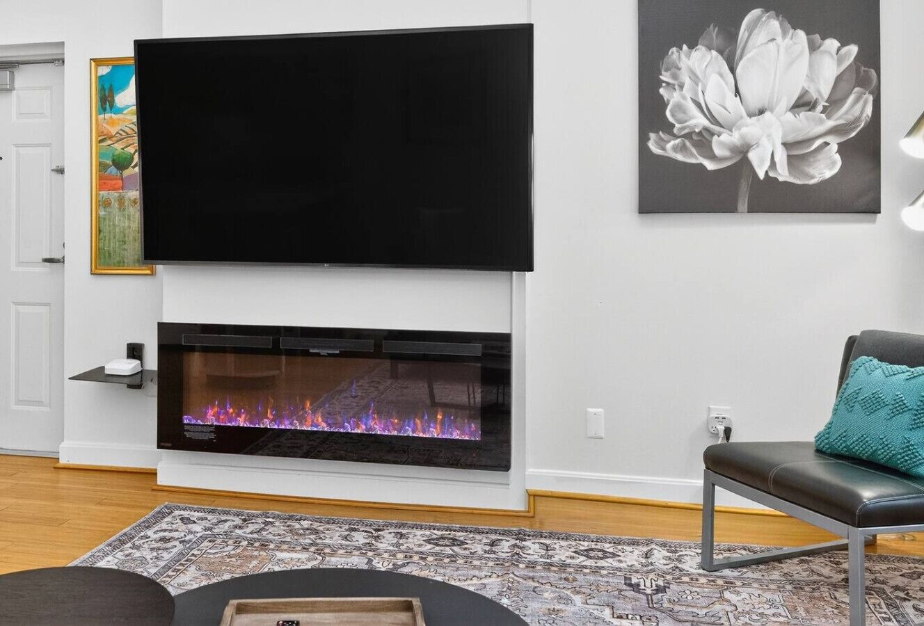 Ein Wohnzimmer mit Kamin, einem flachbildfernseher darüber, einem gemusterten Teppich und einem türkis-farbenen Kissen auf einem schwarzen Sessel.
