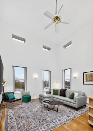 A spacious living room with a high ceiling, a ceiling fan, large windows, a gray sofa, a coffee table, and a patterned rug. - Renovated FL3| Near UC+TQL Stadium | Free Parking (Cincinnati)