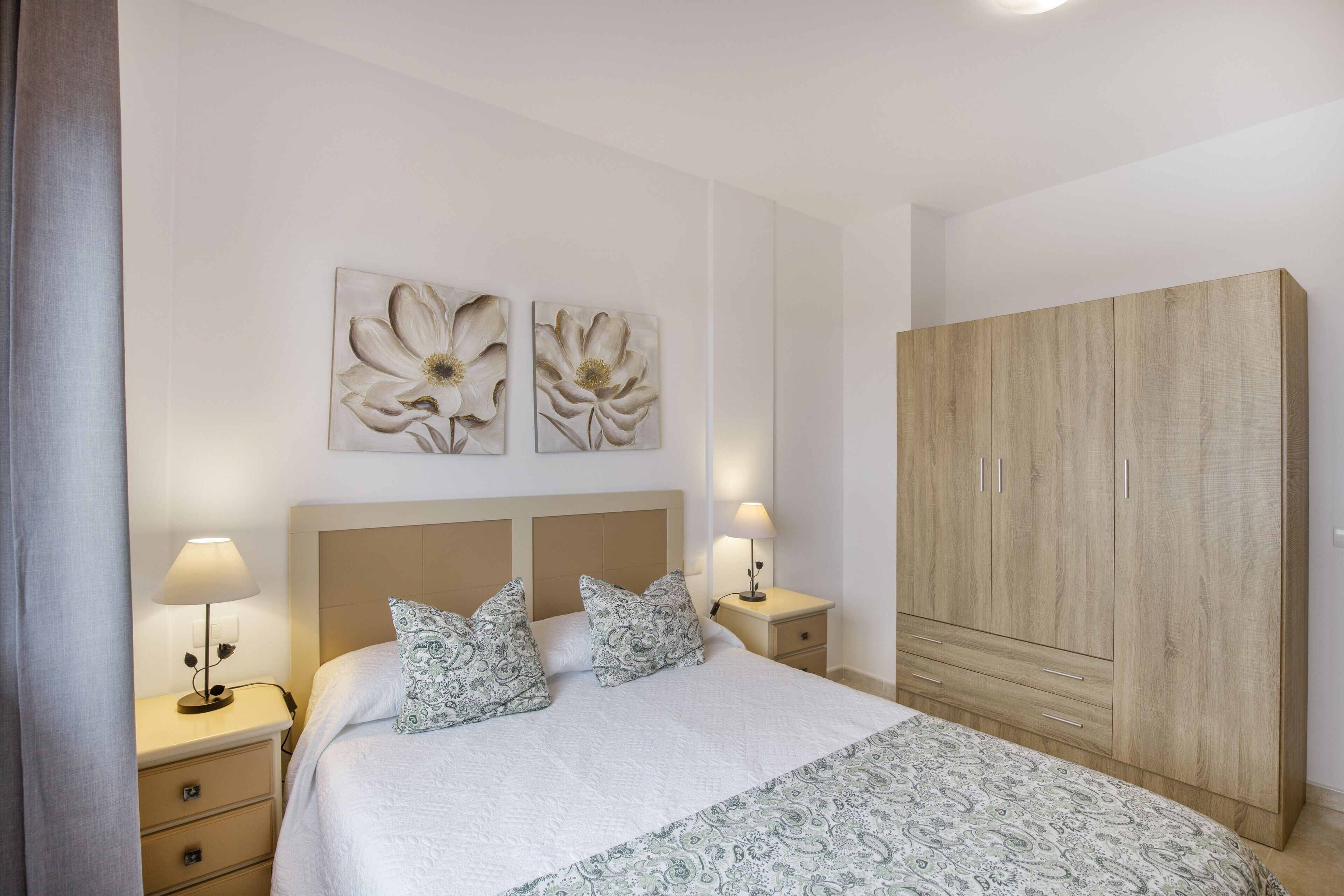 A bedroom with a bed, two wall-mounted lamps, a nightstand, a wooden wardrobe, and two wall art pieces.