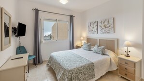 Ein Schlafzimmer mit Bett, Nachttischen, Schreibtisch, Stuhl, Fernseher und einem Fenster mit Vorhängen.