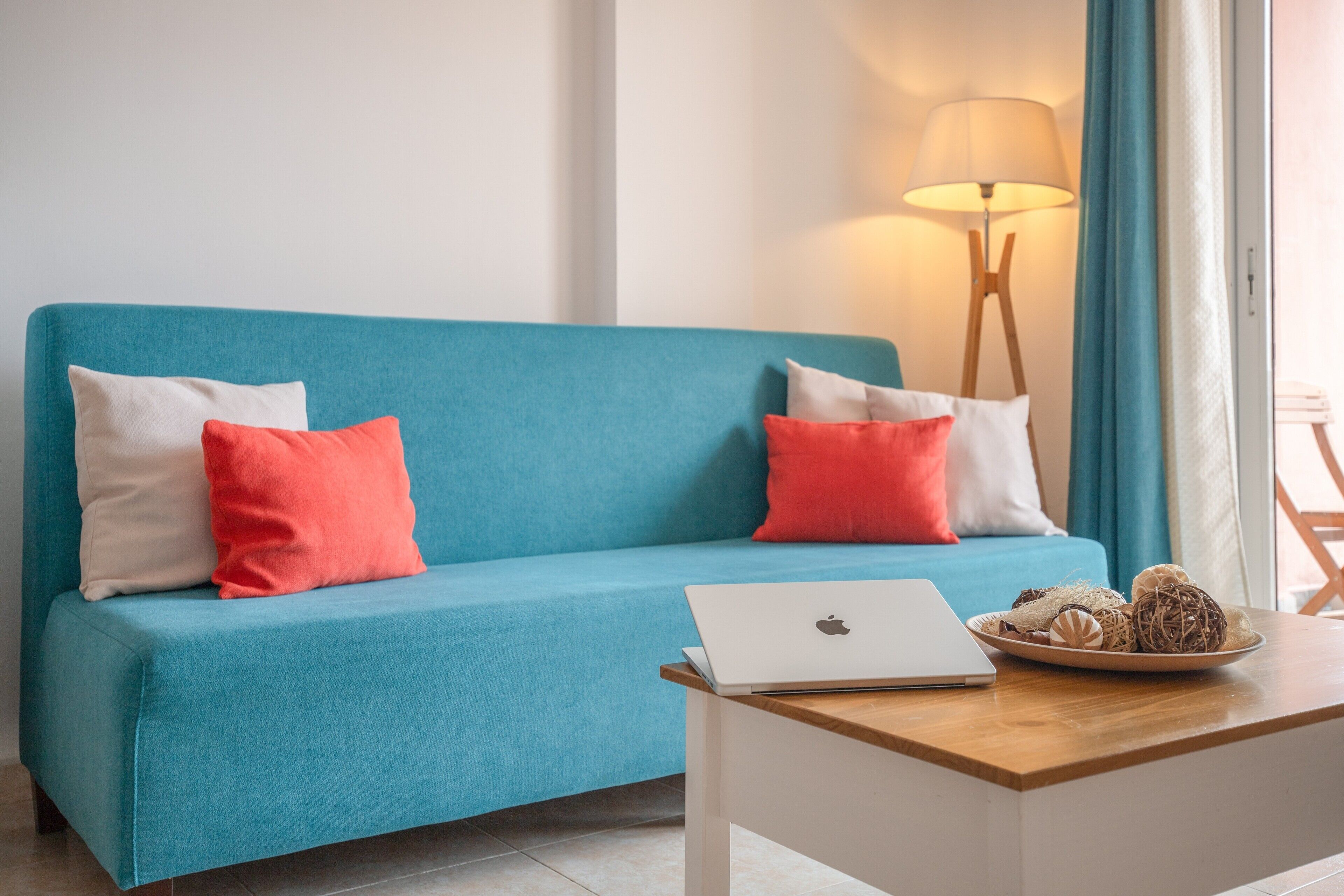Ein türkisfarbenes Sofa mit weißen und roten Kissen, ein Holztisch mit einem Laptop und Dekoration, sowie eine Stehlampe in einem modernen Zimmer.