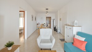 Ein modernes Wohnzimmer mit einem weißen Sessel, einem türkisblauen Sofa mit rotem Kissen, einem Holztisch und einer Küche im Hintergrund.