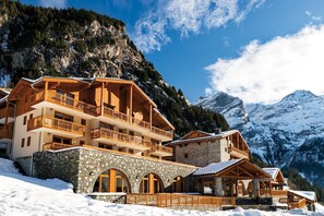 A multi-story chalet with stone foundations and wooden balconies, set against a snowy mountain backdrop. - Appart Ski aux Pieds | Terrasse + Accès piscine (Pralognan-La-Vanoise)