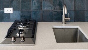 A kitchen with a gas stove, stainless steel sink, dark blue tiled backsplash, wooden shelf with glasses and a vase, and a power outlet.