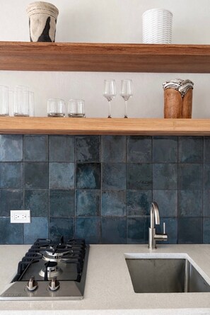 Una cocina con estufa a gas, pileta de acero inoxidable, salpicadero de azulejos azul oscuro, repisa de madera con vasos y un florero, y un enchufe.