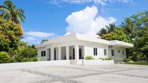 A modern white building with a covered patio area, surrounded by lush greenery and palm trees.