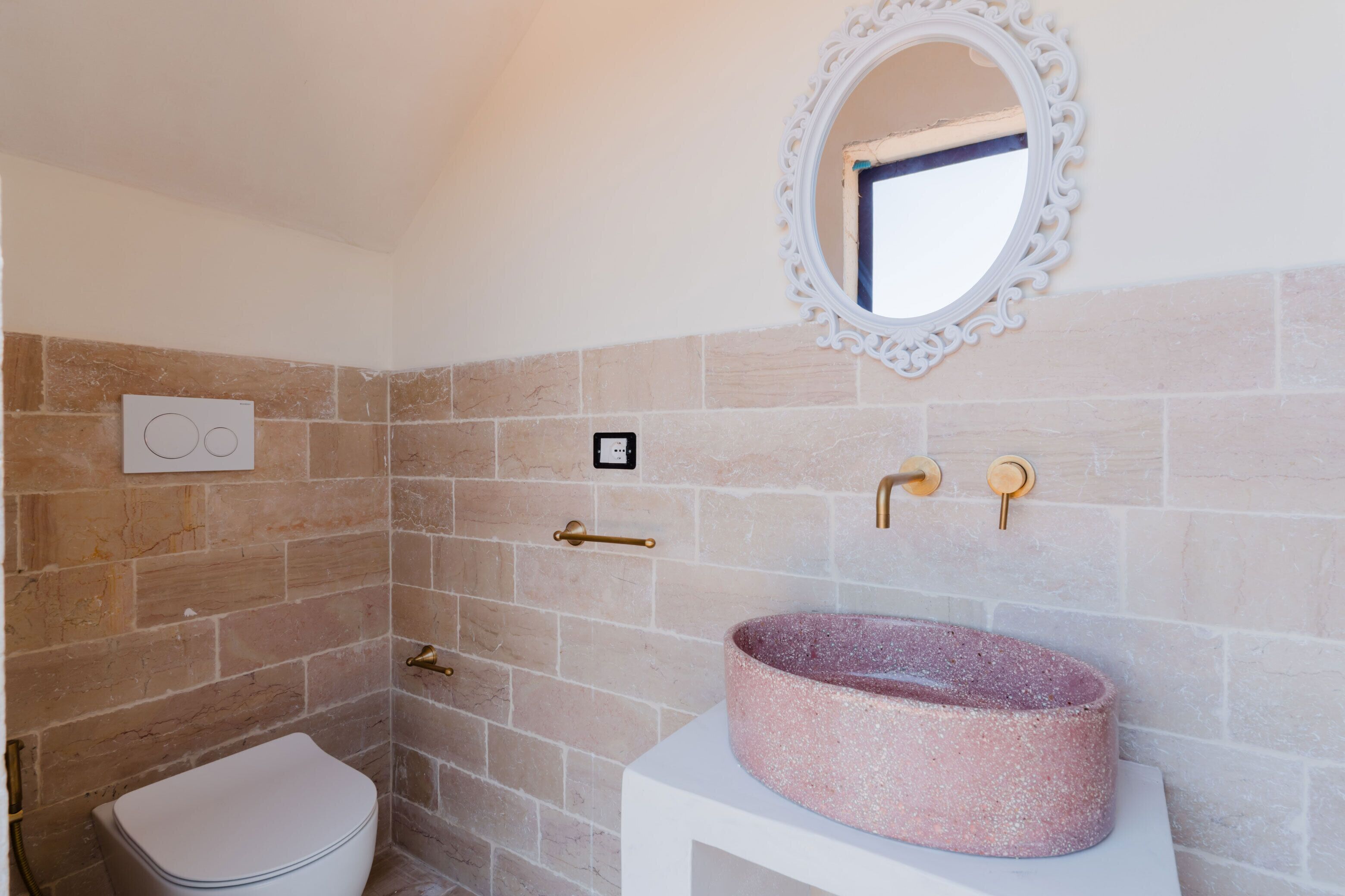 Un baño con un lavabo rosa, un inodoro blanco, una ducha con pared de azulejos color beige y un espejo decorativo.