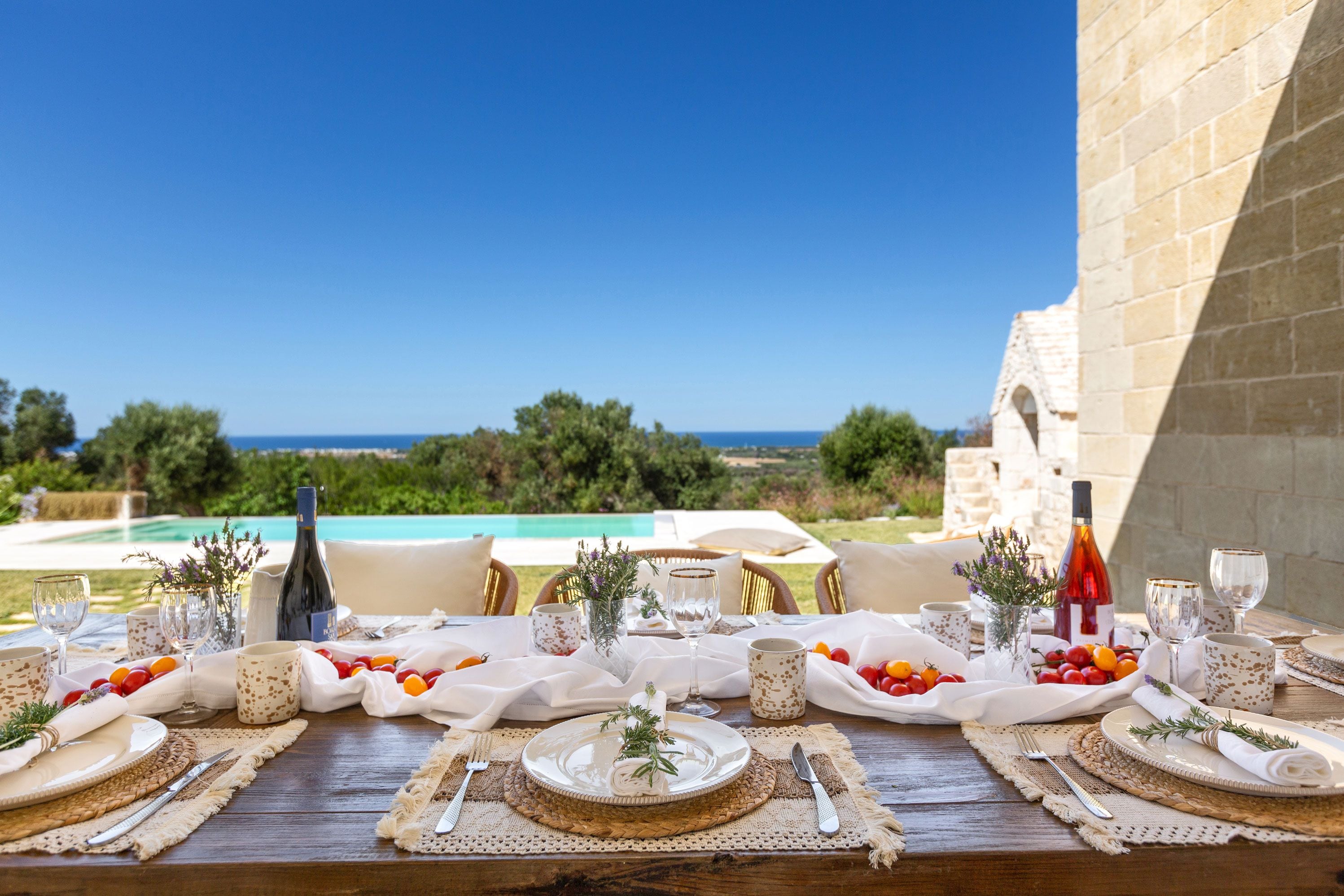 An outdoor dining table set with plates, glasses, and a bottle, overlooking a pool and greenery.