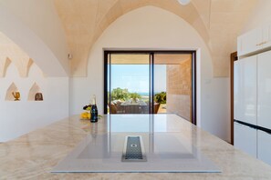 A modern kitchen with a central island, a view to an outdoor dining area, and a large window. - Tenuta Romance by Perle di Puglia (Carovigno)