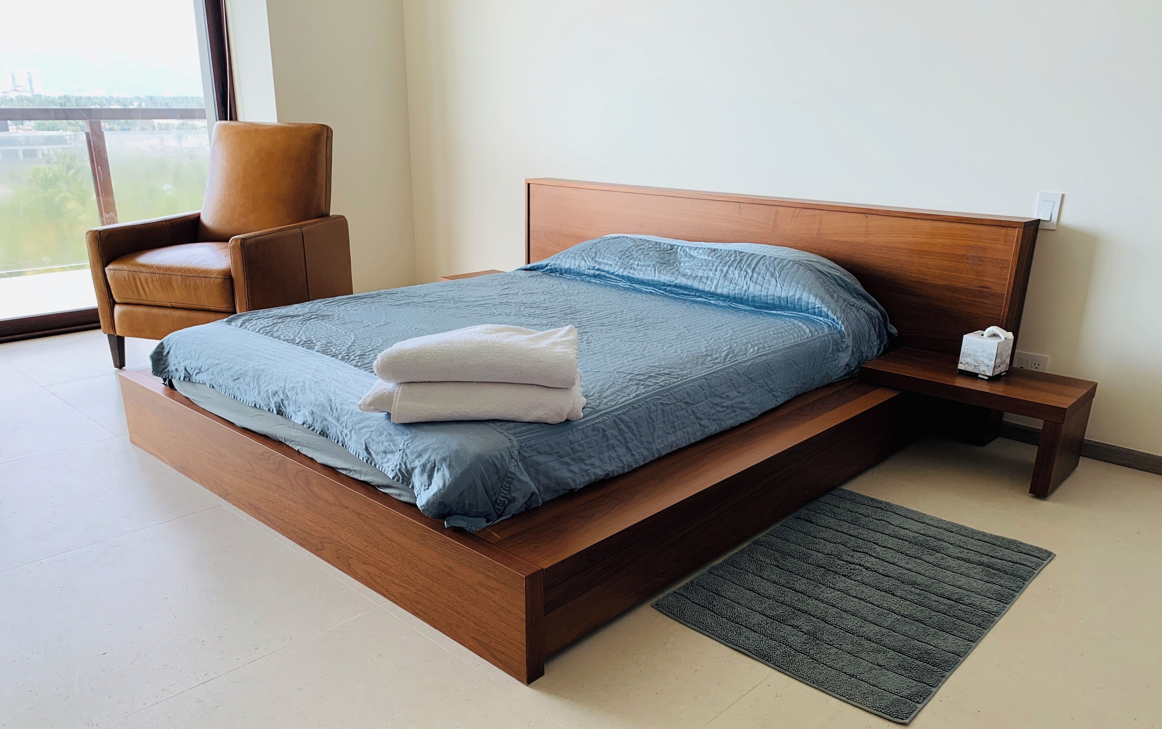 A bedroom with a wooden bed, a nightstand, a chair, and a view of the outdoors.