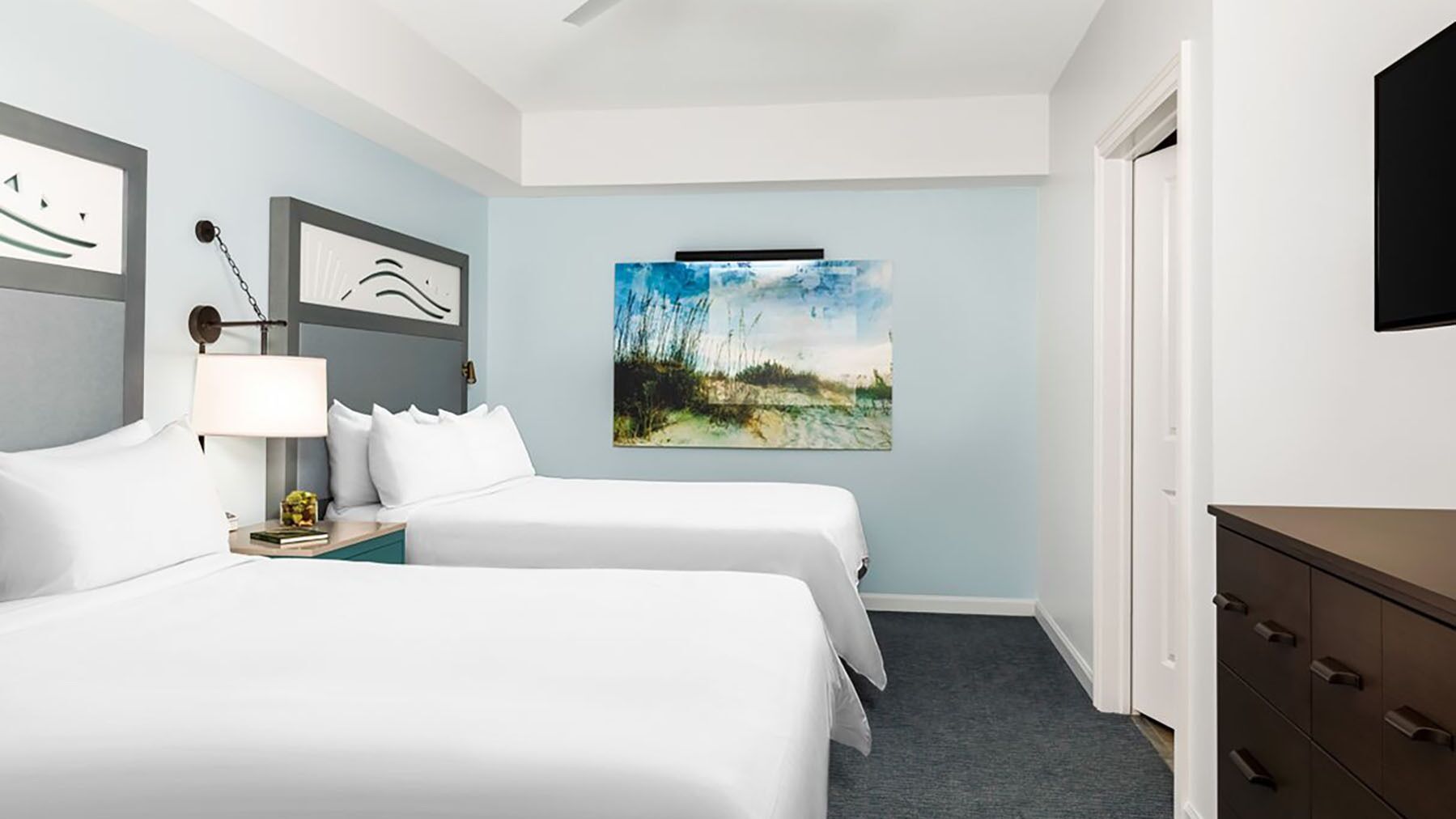 A hotel room with two beds, a desk, a chair, a lamp, and artwork on the walls.