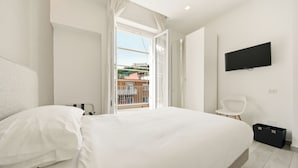 A bedroom with a large bed, a TV mounted on the wall, a balcony with a view of buildings, and a small white chair. - Alpha Domus 15 (Naples)
